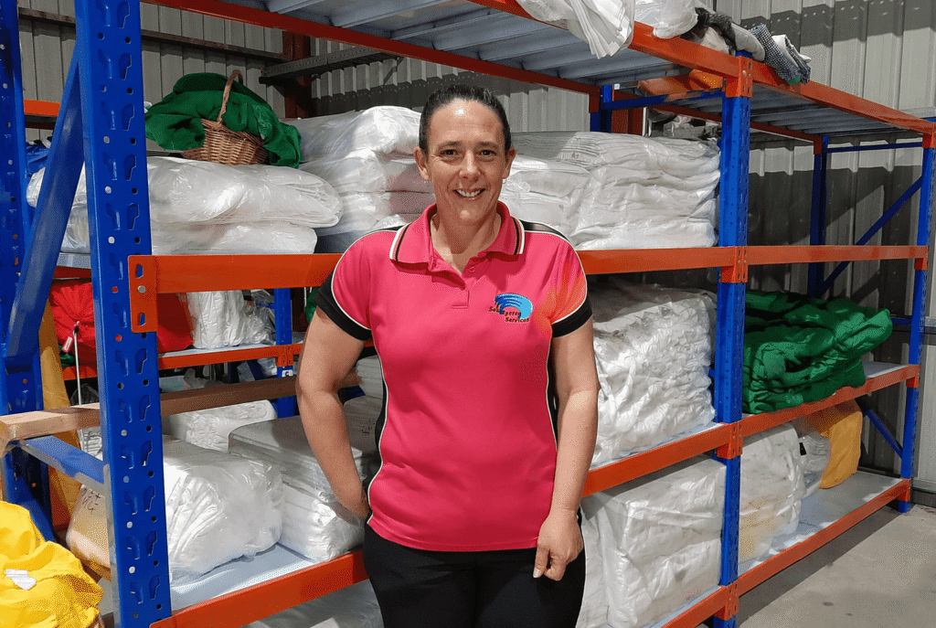 Woman with dark hair pulled back wearing a pink short sleeved t-shirt and black pants standing in front of a rack of white linen.
